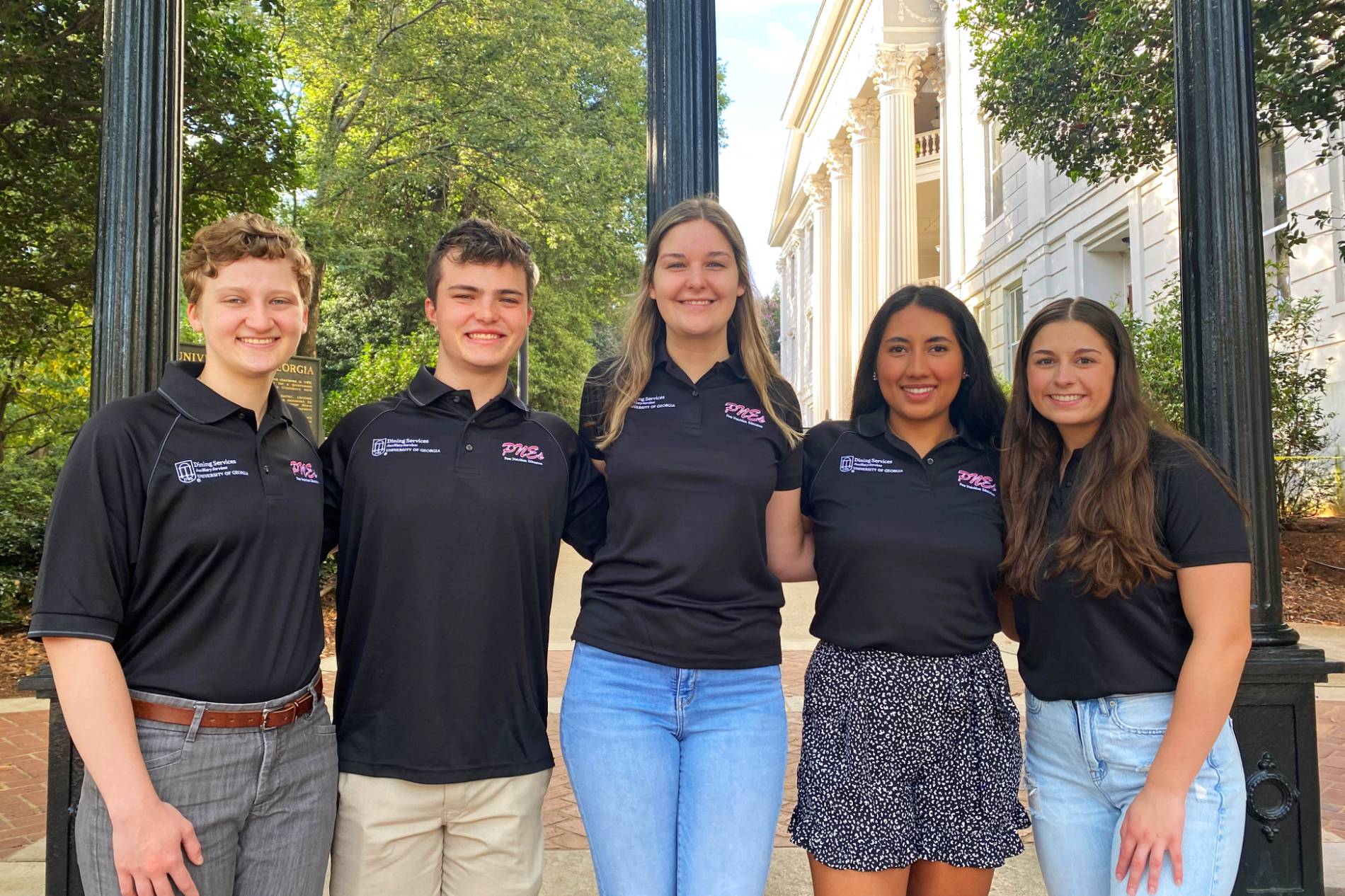 Five students wearing matching black shirts with the acronym "PNEs" stand in front of the UGA Arch and smile at the camera.